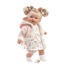 A realistic baby doll with vinyl limbs and a soft body, wearing a white floral dress and a pink cardigan, styled with blonde hair in pigtails and a fringe.