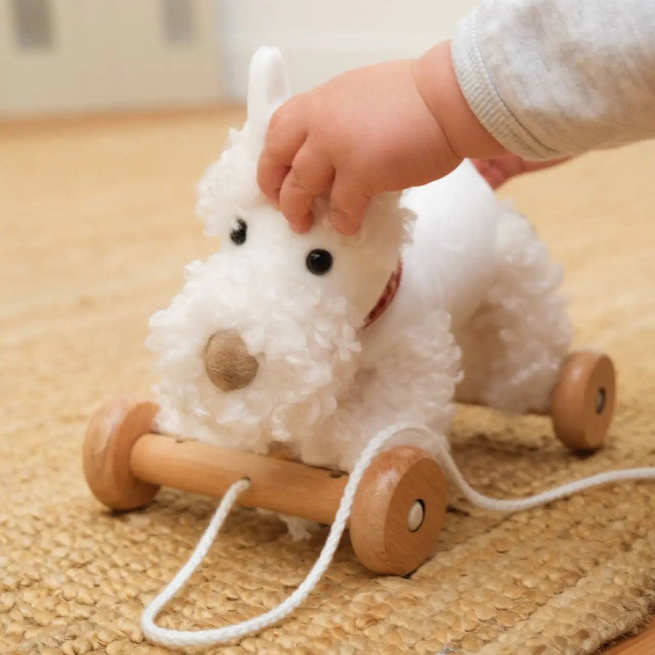 Child's hand pulling a white plush dog toy with wheels on a wooden floor.
