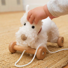 Child's hand pulling a white plush dog toy with wheels on a wooden floor.