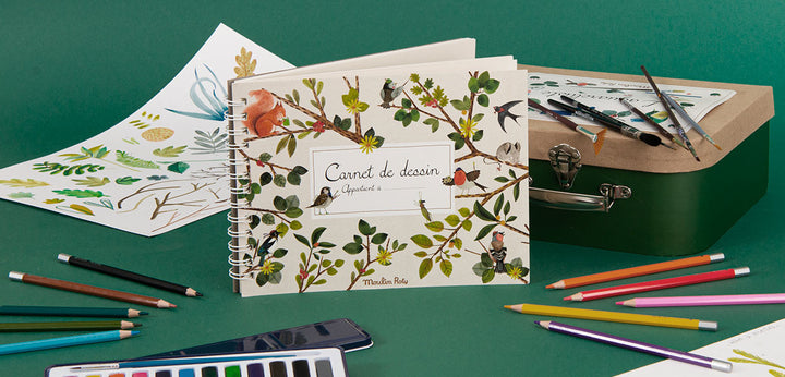 Artistic notebook with nature-themed design on a green surface with art supplies.