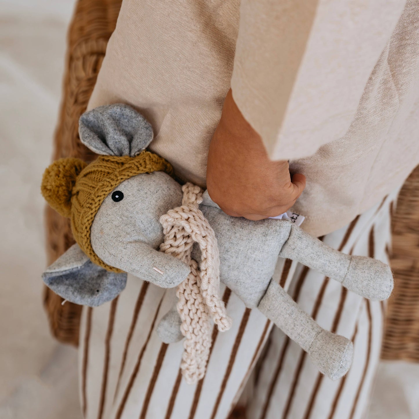 Grey  elephant plush toy with a knitted yellow hat and scarf held by a child wearing a beige coat.