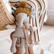 Stuffed elephant toy wearing a knitted hat and scarf in front of a basket with striped fabric.