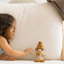 A young child reaches toward a small crocheted plush duck toy wearing a brown knit hat, set against a large cream-colored cushion.