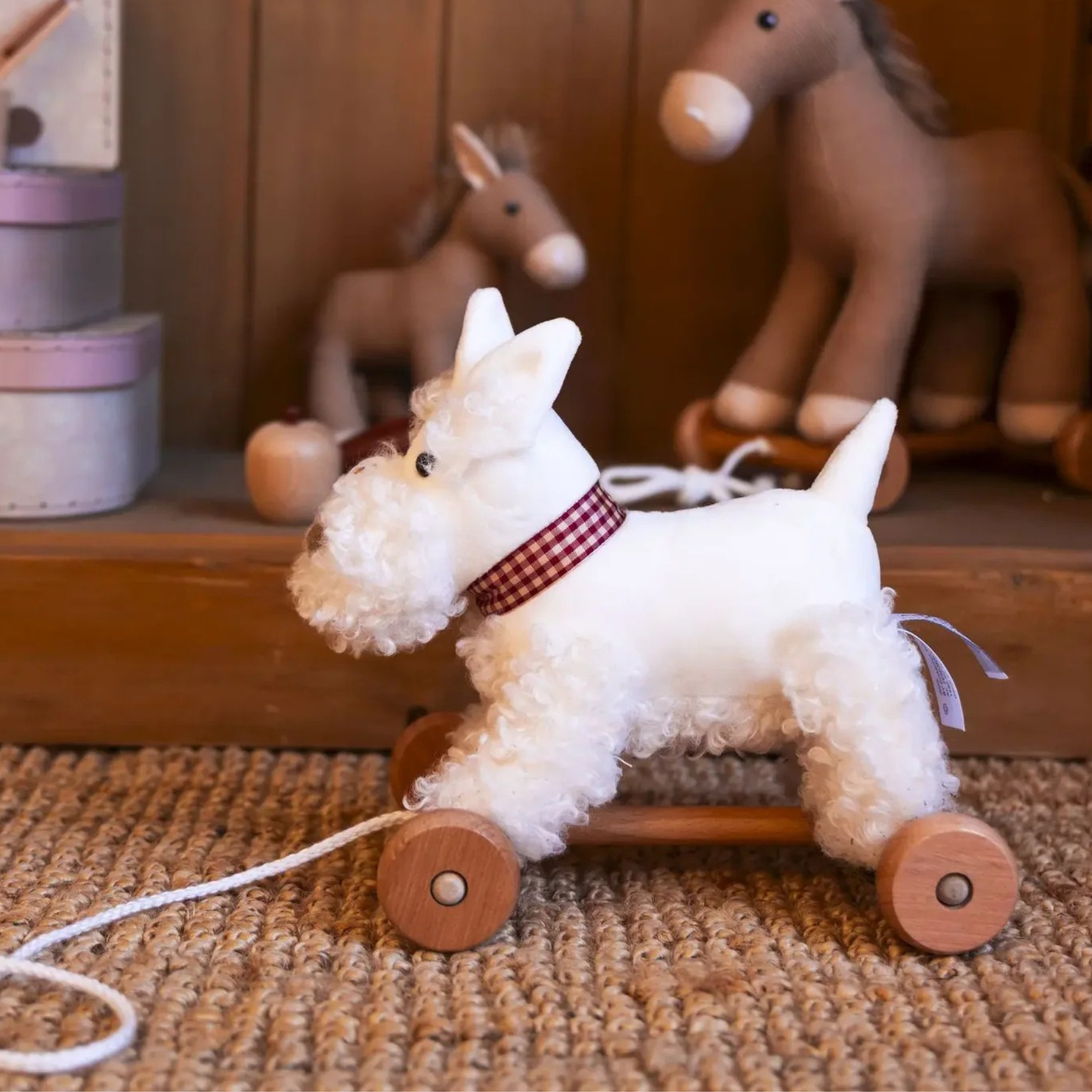 A white plush dog toy on a wooden wheeled pullalong, styled on a woven rug with rustic wooden toys in the background.