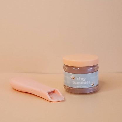 Jar of 'tiny tummies' pretend baby food with a pink spoon on a beige background