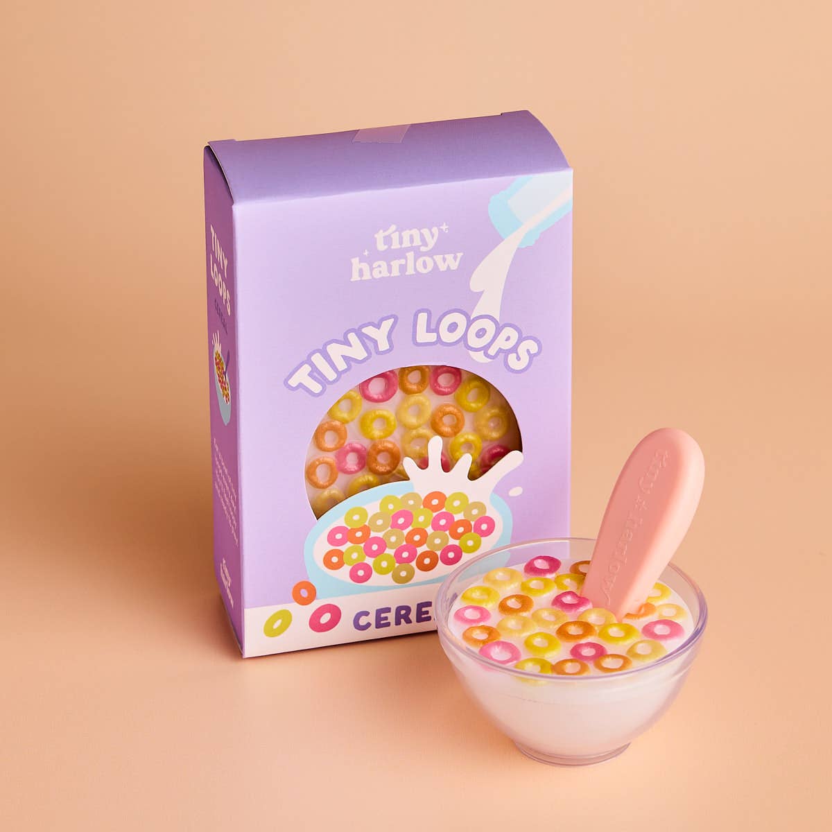 Box of 'Tiny Harlow Tiny Loops' cereal with a bowl of cereal and pink spoon on a beige background