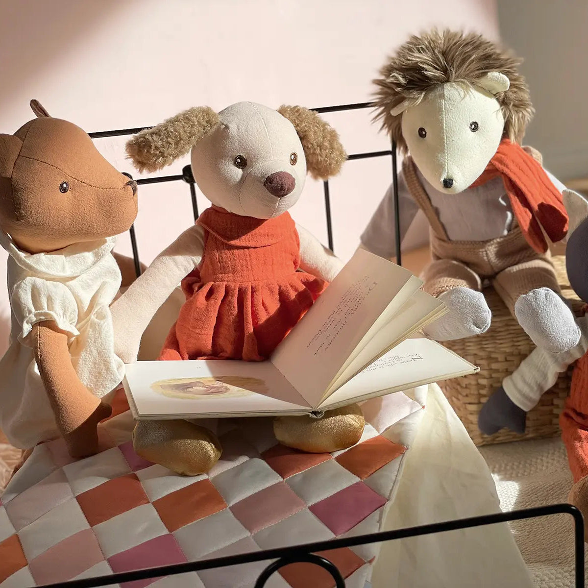 Four stuffed animals sitting on a checkered blanket, with one holding an open book.