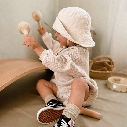 Toddler in a cream romper playing with Love Note Co wooden maracas in a softly lit room.