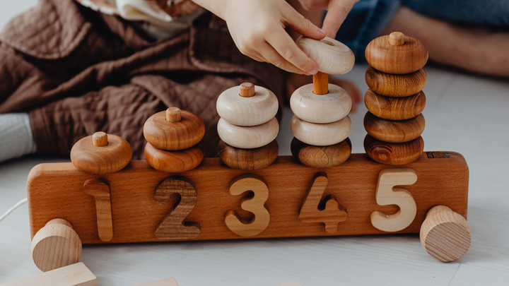 Natural wood roll-along counting toy with numbers 1 to 5 printed on the side. Toy has 5 rods on top, with round stacking discs threaded onto rods.