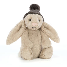 A beige plush toy in the shape of a bunny with a charcoal hat, biscuity paws, and a pastel pink nose, designed for children.