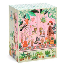 A decorative music box with a floral and garden motif, featuring illustrations of birds, plants, and a young girl. The music box is closed and appears to be made of wood with a paper or fabric decoration.