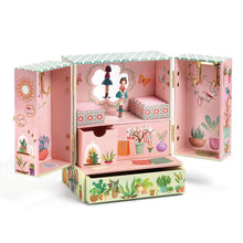 A decorative music box with a floral and garden motif, featuring illustrations of birds, plants, and a young girl. The music box is closed and appears to be made of wood with a paper or fabric decoration.