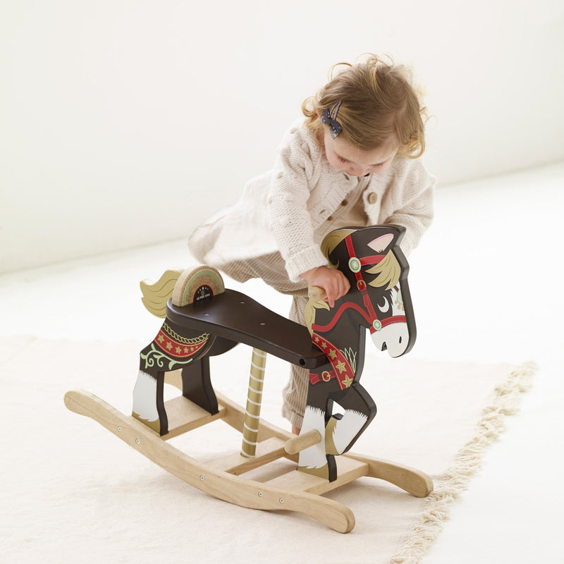 A young child hopping onto a traditional wooden rocking horse with brown and white color scheme, featuring hand and foot grips, and decorative carousel horse and reins artwork.