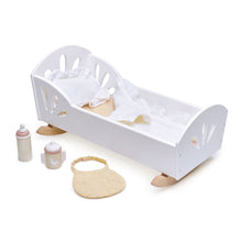 A white wooden dolls cradle pictured on a white background with assorted wooden and fabric doll accessories.