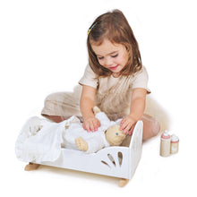 A young child playing with a white doll in a cot-like bed with natural wood rockers. The set includes a blush mattress, muslin blankie, pillow, wooden baby bottle, baby cup, and muslin bib.